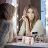 A woman applies makeup in front of a mirror with a portable neoprene cosmetic organizer bag on the counter.