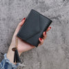 A hand holds a black PU leather coin clutch wallet with a gold chain tassel against a gray background.
