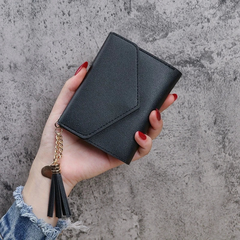 A hand holds a black PU leather coin clutch wallet with a gold chain tassel against a gray background.