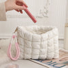 A hand holds a pink quilted puffer makeup bag with a gold chain against a white fluffy background.
