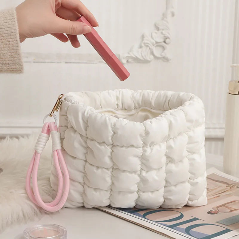 A hand holds a pink quilted puffer makeup bag with a gold chain against a white fluffy background.