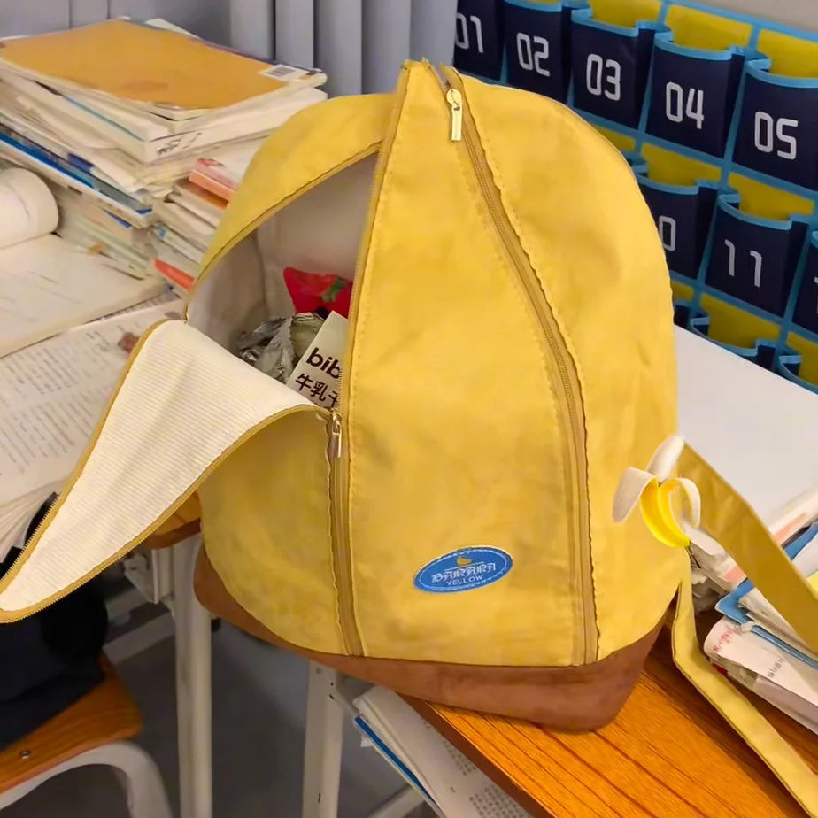 A yellow banana-shaped backpack is shown resting on a wooden surface next to stacked papers and other items.