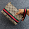 Beige und braun gemustertes Clutch-Handtasche mit rotem und grünem gestreiftem Lederriemen, gehalten von einer Hand.