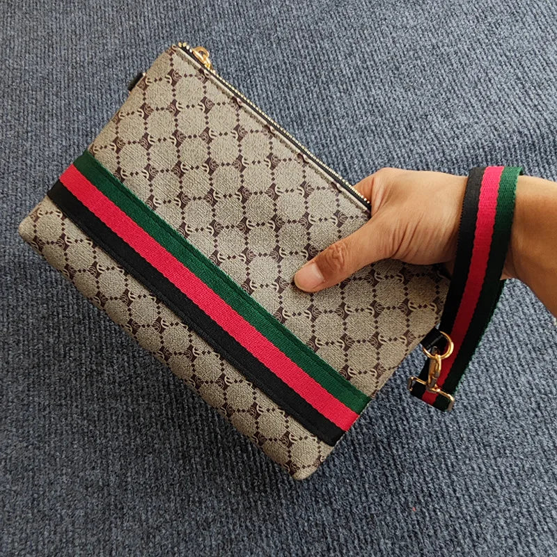Beige und braun gemustertes Clutch-Handtasche mit rotem und grünem gestreiftem Lederriemen, gehalten von einer Hand.