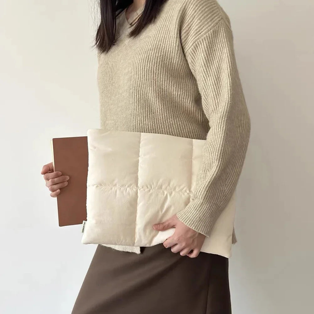 Une personne tient une housse de laptop rembourrée beige contre un pull beige clair et une jupe marron.