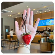A hand holds a red strawberry bag charm keychain against a blurred background of an indoor cafe.
