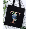 Black canvas tote bag featuring a large gold letter T with a blue floral print against a white background.