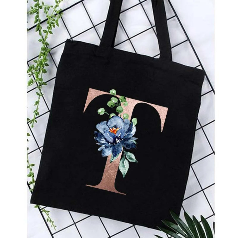 Black canvas tote bag featuring a large gold letter T with a blue floral print against a white background.