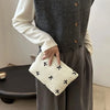 A hand holds a white embroidered makeup pouch against a gray knitted sweater and dark trousers.
