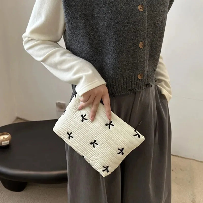 A hand holds a white embroidered makeup pouch against a gray knitted sweater and dark trousers.