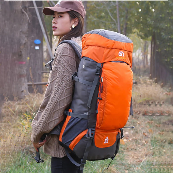 A woman is holding an orange and gray waterproof outdoor climbing backpack in a natural, wooded setting.