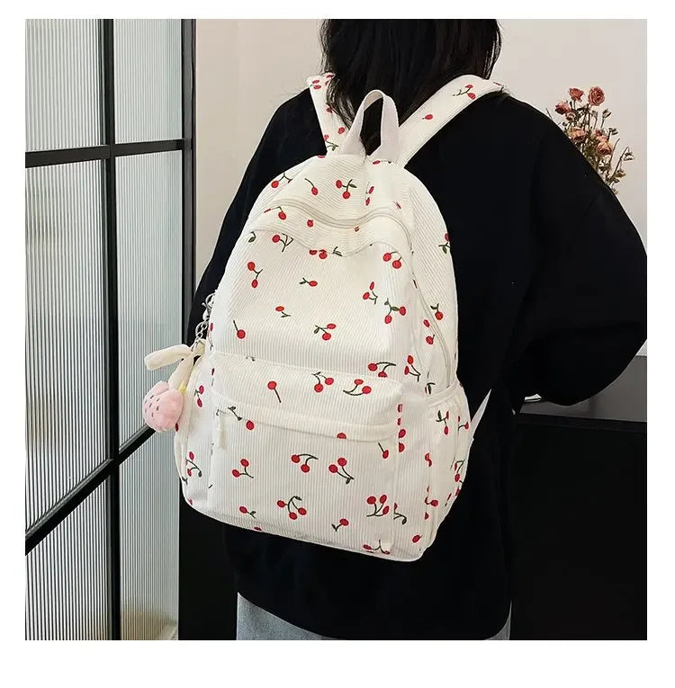A white corduroy fashion backpack with a floral pattern and adjustable strap is worn by a person against a glass door.