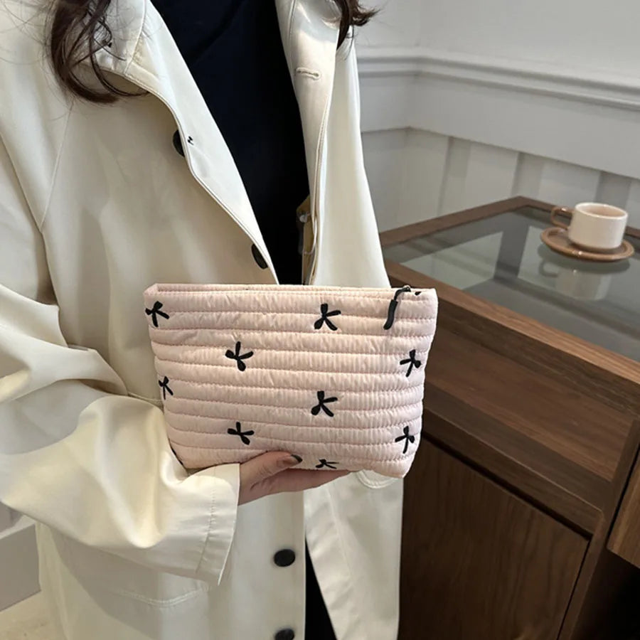 A light pink quilted makeup pouch with black embroidered crosses is held by a person in a white coat.