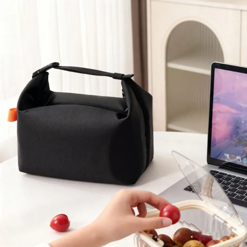 Une main tient une tote bag isotherme noire à côté d'un ordinateur portable et de fruits frais.