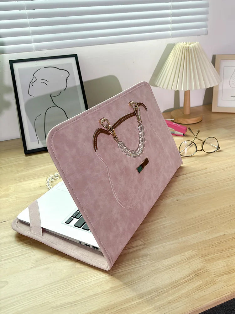 A pink PU laptop sleeve with a leather strap and silver chain is displayed on a wooden desk.
