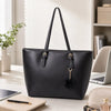 Bolso tote de cuero PU negro con cadena dorada y adorno pompom negro sobre una superficie de madera clara.