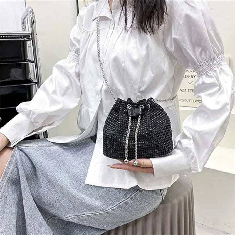 Una mujer modela un bolso cruz de cordón con pedrería negra brillante y cadena plateada contra una camisa blanca y falda gris