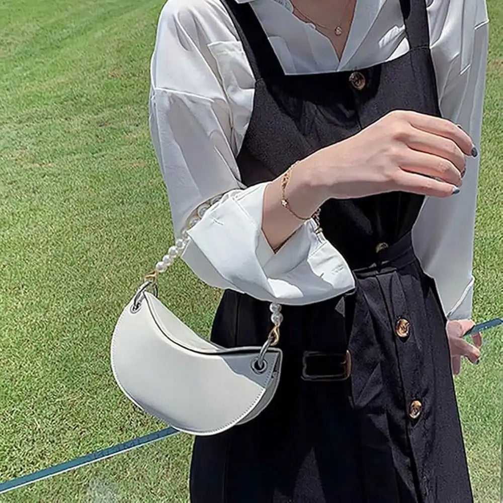 A woman wearing a black apron and white shirt holds a light gray leather handbag with a pearl strap.