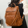 A brown vintage tassel backpack is shown from the back, carried by a person wearing a black dress and white sleeve.