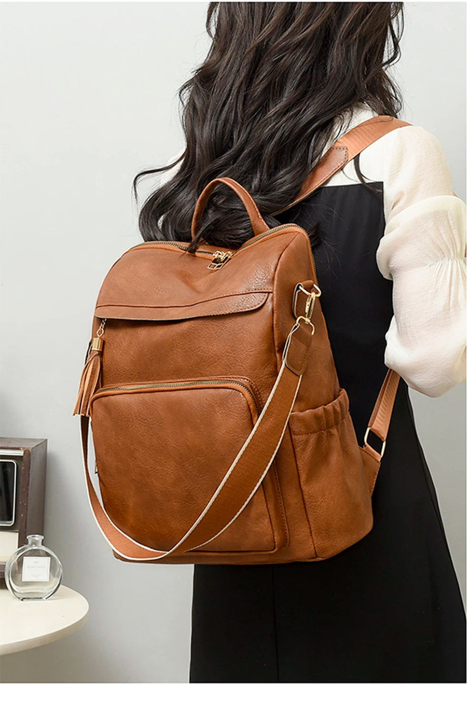 A brown vintage tassel backpack is shown from the back, carried by a person wearing a black dress and white sleeve.