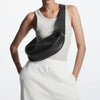 Una mujer modela un bolso cruz de cuero negro genuino con un top de tirantes gris claro y pantalones blancos.