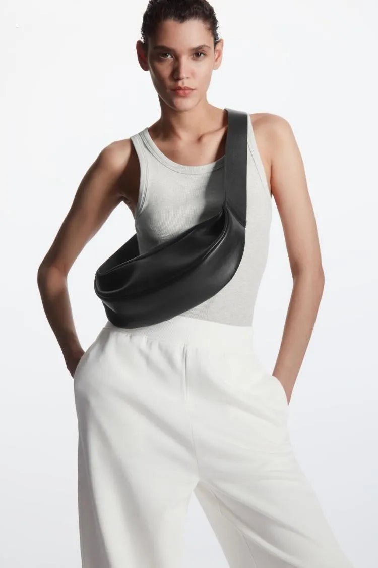 Una mujer modela un bolso cruz de cuero negro genuino con un top de tirantes gris claro y pantalones blancos.