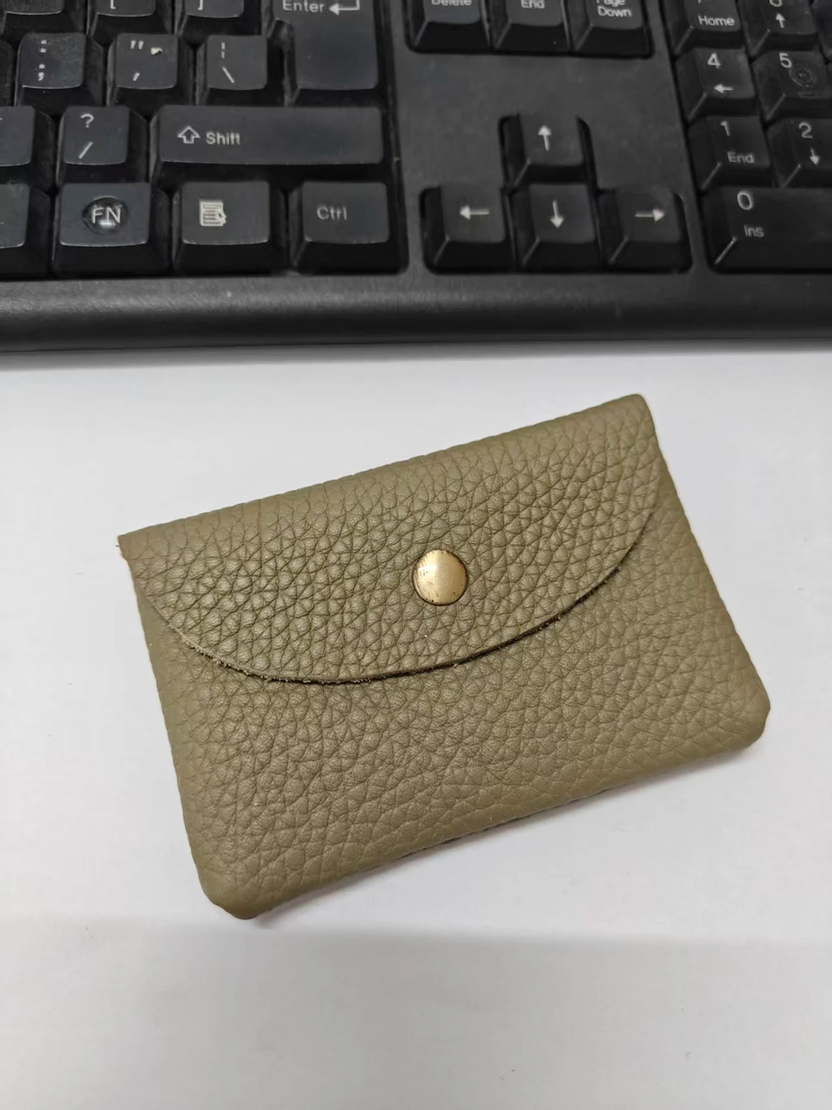 A tan genuine leather card holder with a stitched texture and a round brass stud, placed on a white surface.