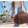 A person wearing jeans is carrying a brown large capacity laptop backpack while standing near a large outdoor structure.