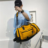 A person wearing a black cap and light blue shirt holds a yellow tactical backpack.