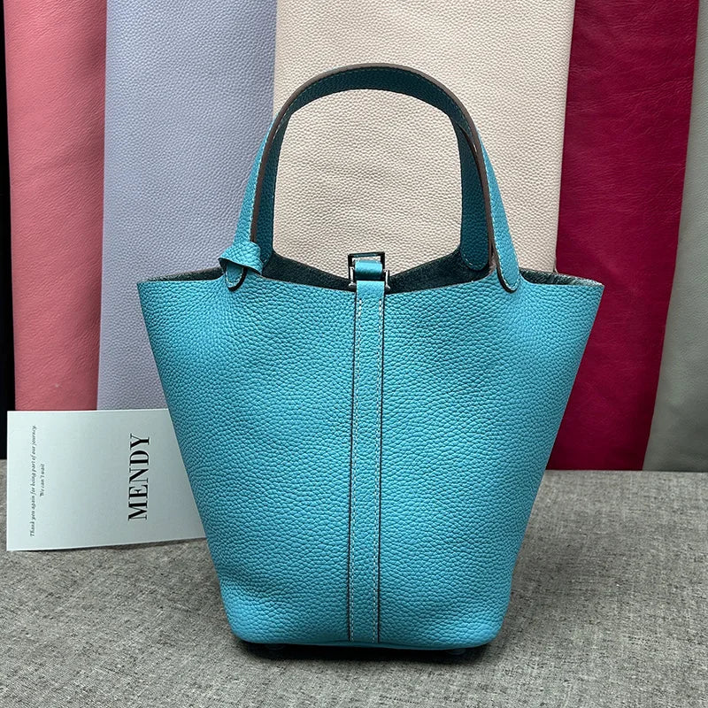 A teal leather bucket bag with a dark gray strap is displayed against a backdrop of various colored fabric panels.