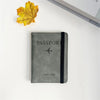A gray passport wallet lies next to a yellow autumn leaf on a white surface.