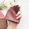 A hand holds a maroon genuine leather zipper card holder and coin purse on a white textured surface.