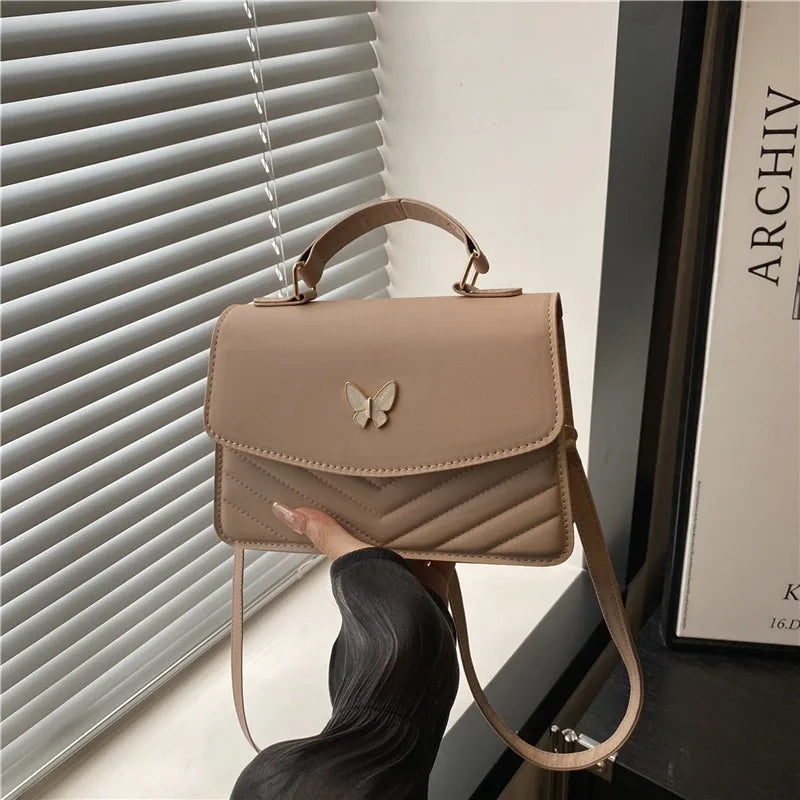 A tan textured shoulder messenger bag is displayed next to a window blind.