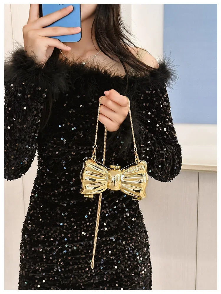 A woman models a gold heart-shaped acrylic evening clutch with a chain strap against a light blue background.