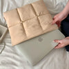 A beige quilted laptop sleeve is placed on a silver laptop in a light-colored bedding setting.