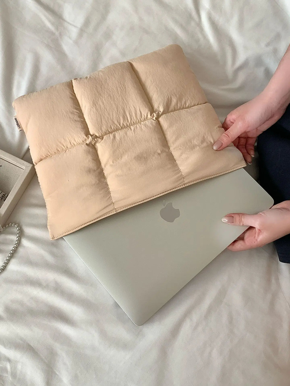 A beige quilted laptop sleeve is placed on a silver laptop in a light-colored bedding setting.