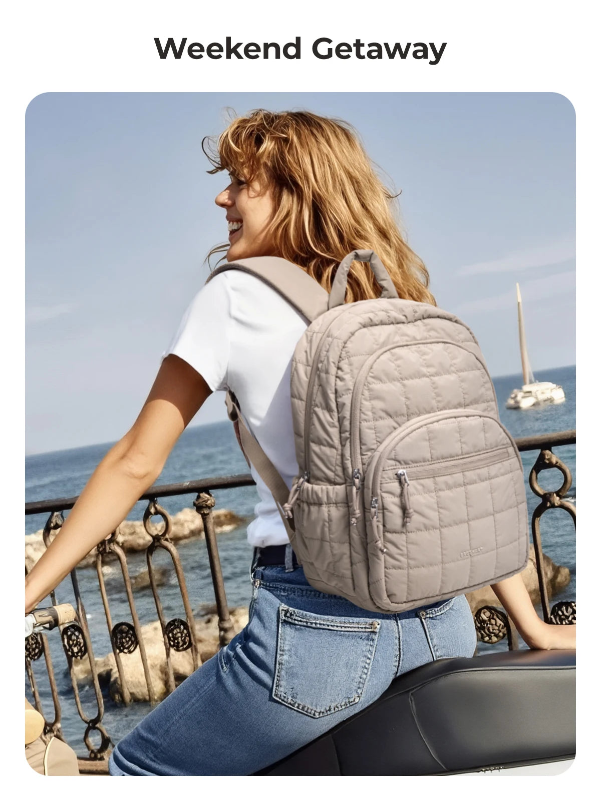 A woman wearing a white shirt and jeans is smiling while carrying a beige quilted casual backpack outdoors by the sea.
