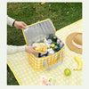 A yellow picnic tote is open, showing various food items and drinks on a checkered blanket outdoors.