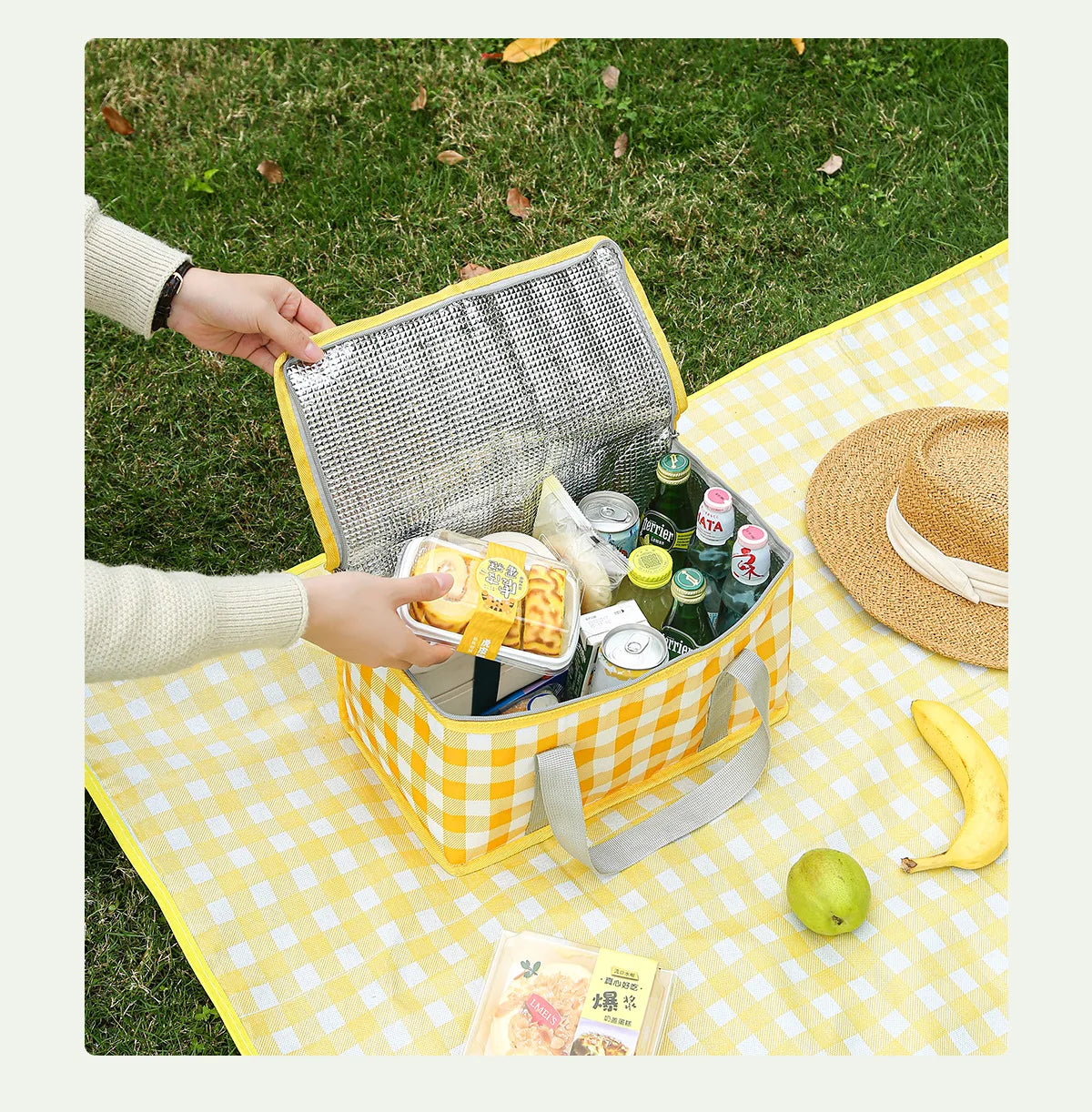 A yellow picnic tote is open, showing various food items and drinks on a checkered blanket outdoors.