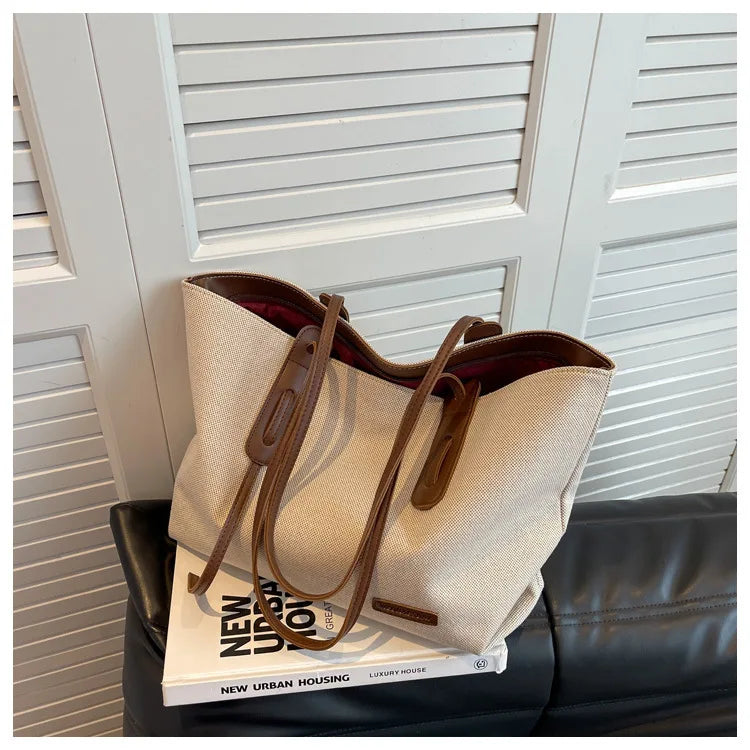 A beige canvas shoulder tote bag with brown leather straps resting on a black leather couch.