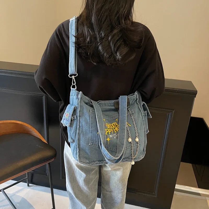 A woman is holding a denim crossbody bag with yellow embroidery against a dark background.