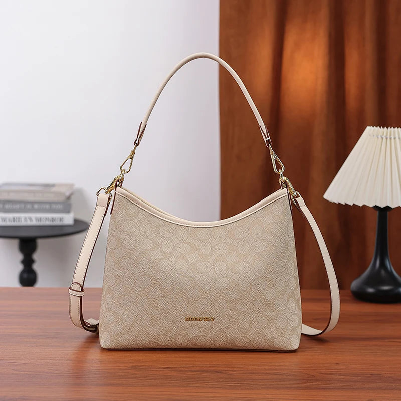 A beige leather crescent tote bag with a letter print and bow accent resting on a wooden table.
