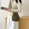 A woman holds a olive green pleated PU leather crossbody bag while wearing a cream sweater and white skirt.