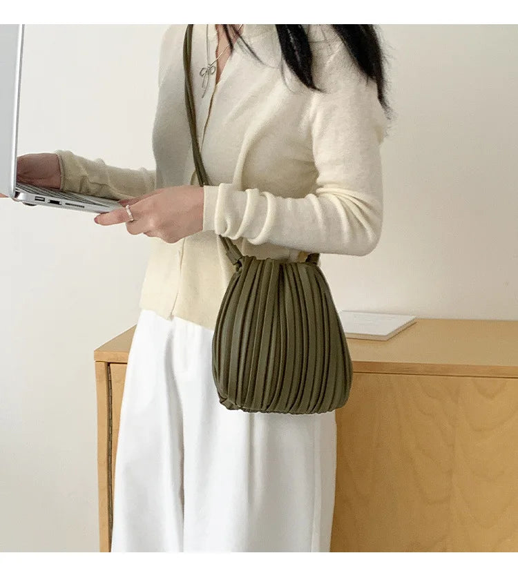 A woman holds a olive green pleated PU leather crossbody bag while wearing a cream sweater and white skirt.