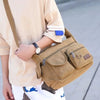A person is holding a tan canvas tool bag with multiple pockets and a strap, next to a light-colored shirt.