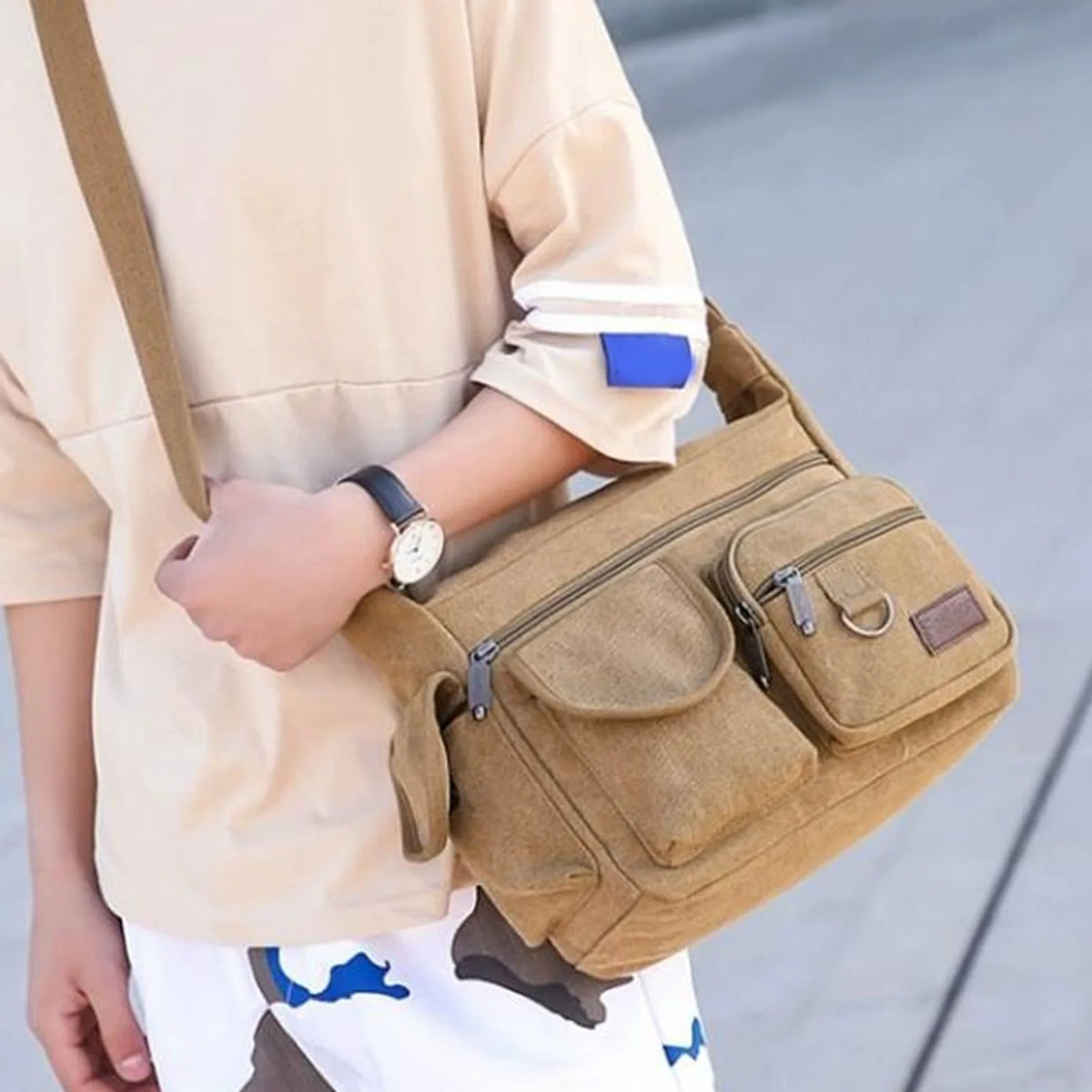 A person is holding a tan canvas tool bag with multiple pockets and a strap, next to a light-colored shirt.