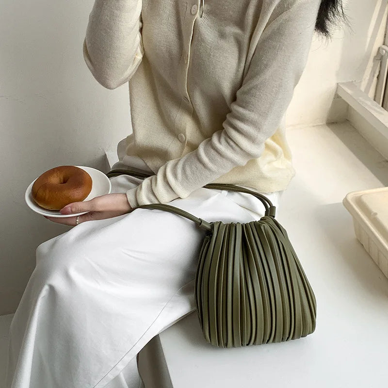 A woman wearing a cream-colored top and white pants holds a olive green PU leather pleated crossbody bag.