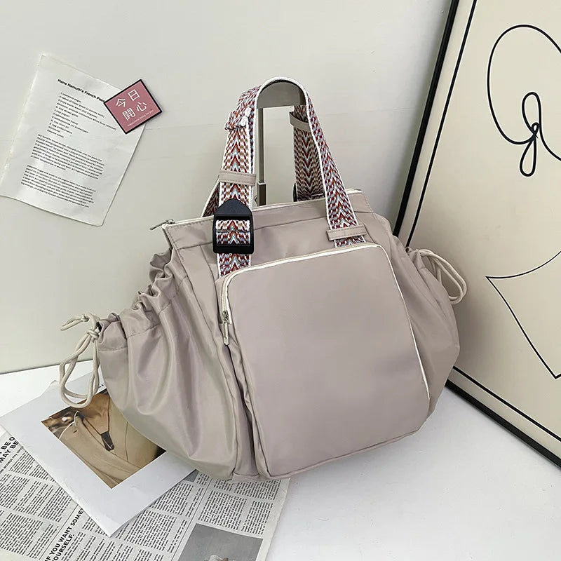 A light beige nylon shoulder bag with striped straps and a flap closure is displayed next to some papers.