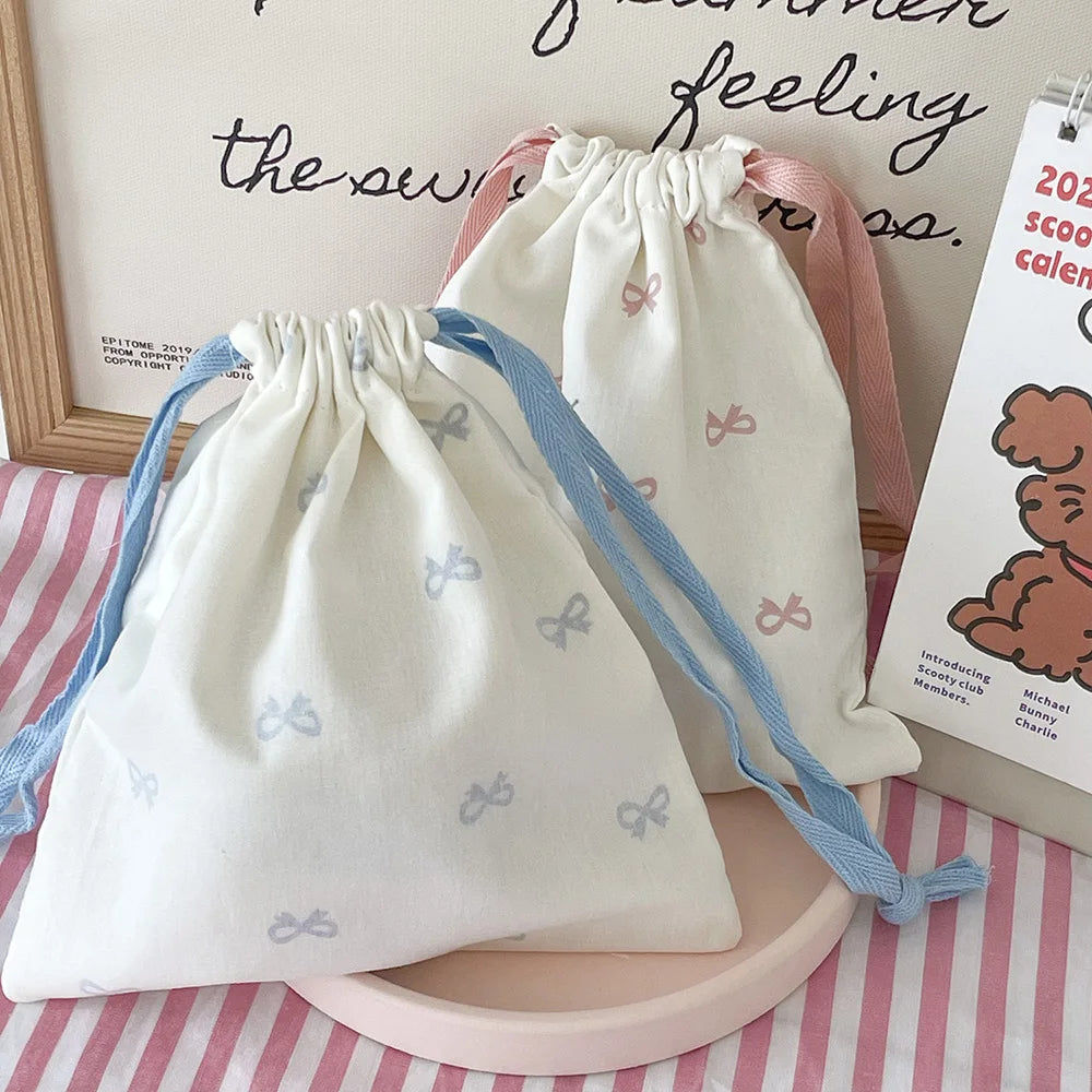 A white canvas makeup bag with pink and blue ribbon ties is displayed on a pink tray.