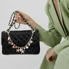 A hand holds a black quilted leather bag with gold chain charms and keychains against a light green blazer.